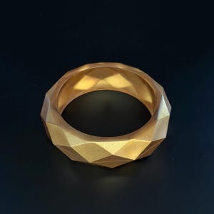 May include: A gold-colored bangle bracelet with a geometric, faceted design. The bracelet has a series of diamond-shaped facets around its circumference. The bangle is made of a solid material and has a smooth, matte finish. The background is black.