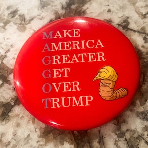 May include: Red button with the text "MAKE AMERICA GREATER GET OVER TRUMP" in white. A cartoon worm with a yellow hairpiece is on the right side of the button.