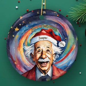 May include: A round, colorful Christmas ornament featuring a portrait of Albert Einstein wearing a Santa hat. The ornament has a swirling galaxy background with stars and the text "Say to the Universe 2024". The name "Jasper" is written on the Santa hat.