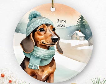 May include: A round Christmas ornament featuring a watercolor illustration of a dachshund wearing a blue hat and scarf. The dog is set against a winter landscape with a house and trees. The name "Jessica" and the year "2025" are written on the ornament.