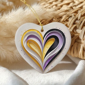May include: A heart-shaped ornament with a layered design in yellow, white, purple, and black. The ornament has a gold string for hanging and is set against a soft, neutral background. The design is a non-binary pride symbol.