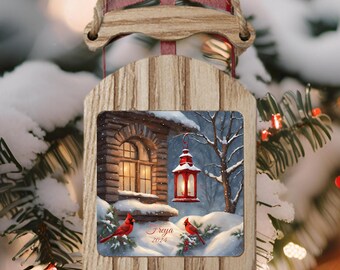 May include: A wooden Christmas ornament shaped like a sled with a winter scene of a cabin with a lit window and a red lantern. Two red cardinals are perched on branches in the snow. The ornament is personalized with the name "Freya" and the year "2024".