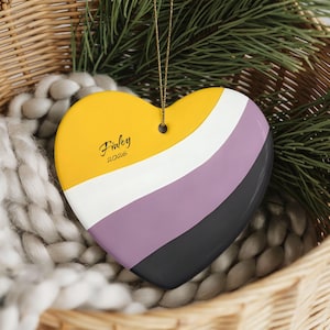 May include: Heart-shaped ornament with a non-binary pride flag design. The ornament features yellow, white, purple, and black stripes. The name "Finley" and the year "2026" are written on the yellow section. The ornament hangs from a gold string and is displayed in a wicker basket.