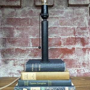 May include: A unique table lamp crafted from a stack of vintage books, including "Moby-Dick" and "Treasure Island." The lamp features a black metal base and a black lamp post with a decorative lightbulb. The background is a red brick wall.