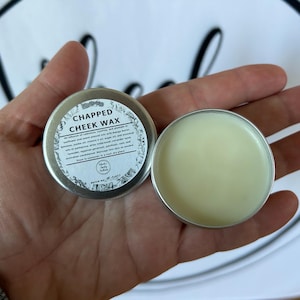 May include: Open tin of Chapped Cheek Wax, a skincare product, held in a hand. The tin is silver with a white label that includes the product name and ingredients. The wax is a light yellow color.