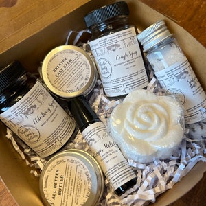 May include: A gift box filled with natural wellness products, including a white bath bomb shaped like a rose, a small bottle of elderberry syrup, a roll-on essential oil blend, a jar of all-butter balm, a jar of breathe easy balm, and a bottle of cough syrup. The box is lined with white shredded paper.
