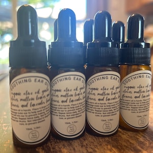 May include: Five small amber glass bottles with dropper tops, each labeled "SOOTHING EAR OIL" and containing organic olive oil, gold olives, mullein leaves, lavender flowers, and lavender buds. The bottles are arranged in a row on a wooden surface.