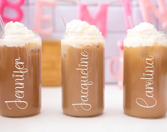 May include: Three clear glass tumblers filled with iced coffee, topped with whipped cream, and a clear straw. Each glass has a name in white script: Jennifer, Jacqueline, and Carolina. The background includes pink and white decor.