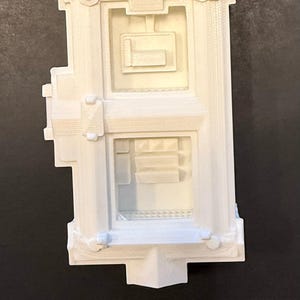 May include: A white architectural model with rectangular windows and decorative features. The model is approximately 7.6 cm tall and is placed next to a ruler for scale.