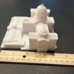 May include: A white 3D printed model of a building with a dome on top. The model is about 4 inches long and 2 inches wide. It is sitting on a wooden ruler.