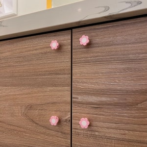 May include: Close-up of two cabinet doors with pink flower-shaped knobs. The cabinet doors are made of wood with a light brown finish.