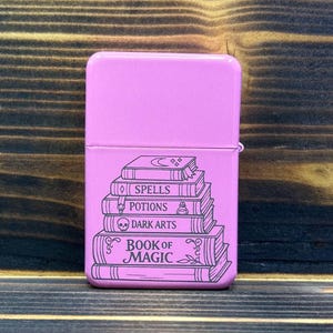 Op de afbeelding: Een rechthoekige roze metalen aansteker met een zwarte lijntekening van een stapel boeken. De boeken zijn voorzien van de teksten Spells, Potions, Dark Arts en Book of Magic, inclusief een halve maan en een schedelpictogram.