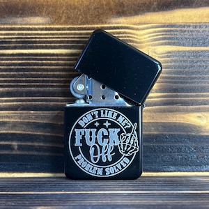 May include: A black metal flip-top lighter with a silver graphic print. The design features a skeleton hand making a middle finger gesture, surrounded by the text "DON'T LIKE ME? FUCK OFF PROBLEM SOLVED" in a circular layout with small stars.