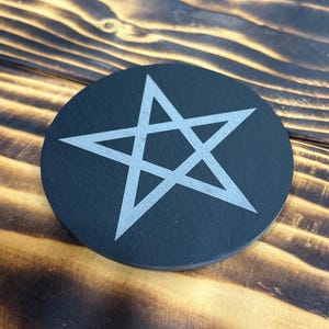 Pode incluir: Um porta-copos circular de ardósia preta com um design de estrela de cinco pontas gravado em cinza claro no centro. O porta-copos tem um acabamento mate suave e uma textura de pedra natural distinta, exibindo um visual geométrico simples.