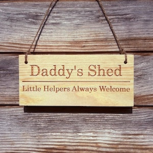 Puede incluir: Un letrero rectangular de madera clara que cuelga de un cordel marrón. El letrero presenta el texto grabado "Daddy's Shed" en la línea superior y "Little Helpers Always Welcome" en la línea inferior, separados por una línea horizontal.