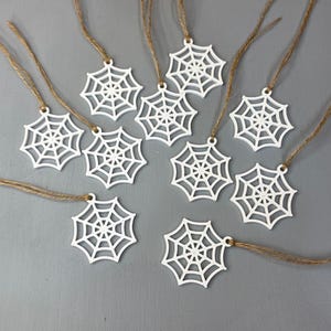 May include: A collection of ten white spider web ornaments, each featuring a die-cut geometric web pattern. Every ornament is attached to a thin, natural brown jute twine loop, allowing for easy hanging. The web designs are uniform in shape.