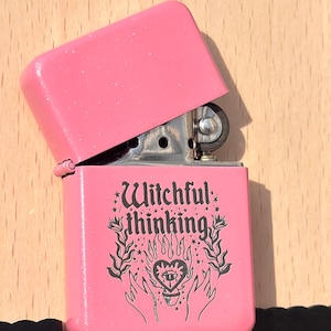 May include: A pink lighter with the top open, revealing the flint wheel. The lighter has the words "Witchful thinking" and a graphic of a heart with an eye, flames, and hands. The lighter is on a wooden surface.