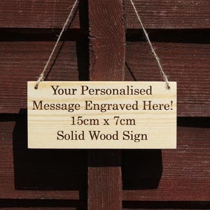 May include: A rectangular light-coloured solid wood sign hanging from a single nail by a thin jute twine string. The sign features the engraved text: Your Personalised Message Engraved Here! 15cm x 7cm Solid Wood Sign. The wood has a natural grain.