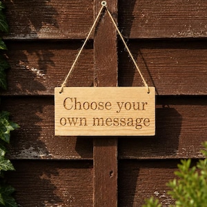 May include: A rectangular light brown wooden sign featuring the engraved text "Choose your own message" in a dark font. The sign is suspended by a thin piece of twine attached to a small metal screw, allowing it to hang vertically.
