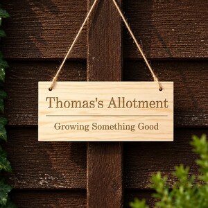 May include: A rectangular light-colored wooden sign with a natural grain, suspended by a thin jute twine. The sign features the engraved text "Thomas's Allotment" above a horizontal line, with the phrase "Growing Something Good" below it.