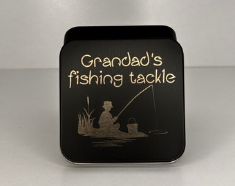 May include: A black metal box with the words "Grandad's fishing tackle" in gold lettering. The box features an etched illustration of a person fishing, with reeds and a bucket. The box is square with rounded corners.