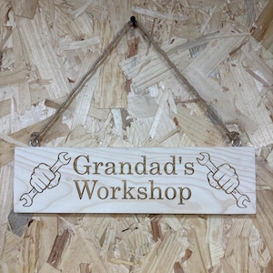 May include: Wooden sign with the text "Grandad's Workshop" and illustrations of hands holding wrenches. The sign is rectangular, light-colored, and hangs from a jute rope. The background is a textured wood panel.