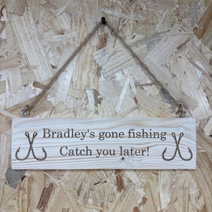 Könnte beinhalten: Helles Holzschild mit dem Text "Bradley's gone fishing Catch you later!" und Angelhaken-Grafiken. Aufgehängt an einem Juteseil und Metallösen. Der Hintergrund ist hellbraunes Holz.