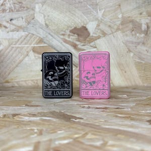 May include: Two rectangular lighters, one black and one pink, each featuring a skull design and the text "THE LOVERS." The black lighter has a silver design, while the pink lighter has a pink design. Both lighters are on a wooden surface.