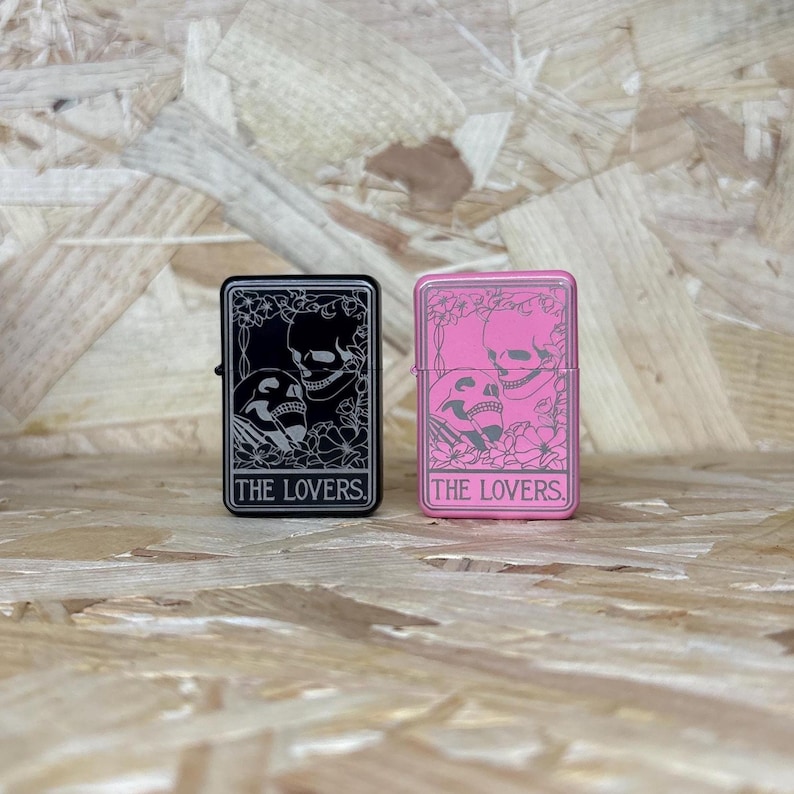 May include: Two rectangular lighters, one black and one pink, each featuring a skull design and the text "THE LOVERS." The black lighter has a silver design, while the pink lighter has a pink design. Both lighters are on a wooden surface.