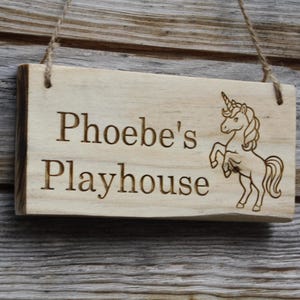 Może przedstawiać: Prostokątna drewniana tablica z napisem "Phoebe's Playhouse" i ilustracją jednorożca. Tablica z jasnego drewna zawieszona jest na sznurkach. Tekst i jednorożec są grawerowane.