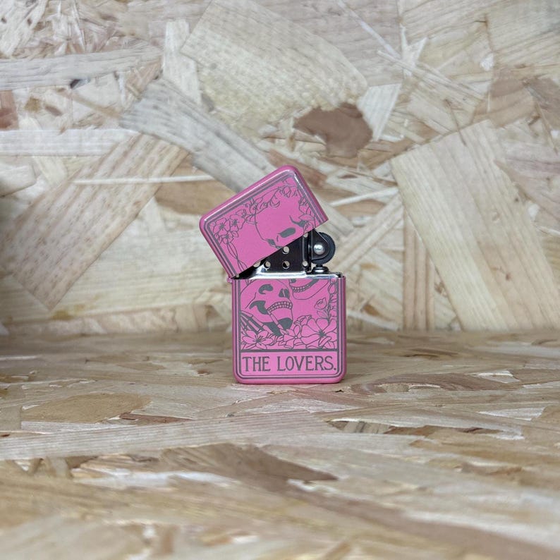 May include: A pink metal lighter with a skull and floral design. The lighter is open, revealing the interior. The text "THE LOVERS" is displayed on the front.