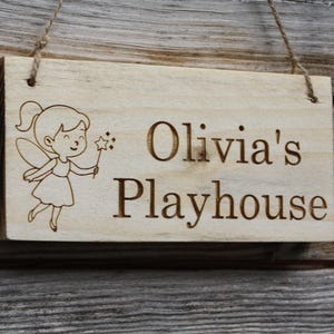 Puede incluir: Cartel de madera rectangular con la inscripción "Olivia's Playhouse" y una ilustración de hada. El cartel tiene un acabado de madera clara y una cuerda para colgar. El hada sostiene una varita en forma de estrella.