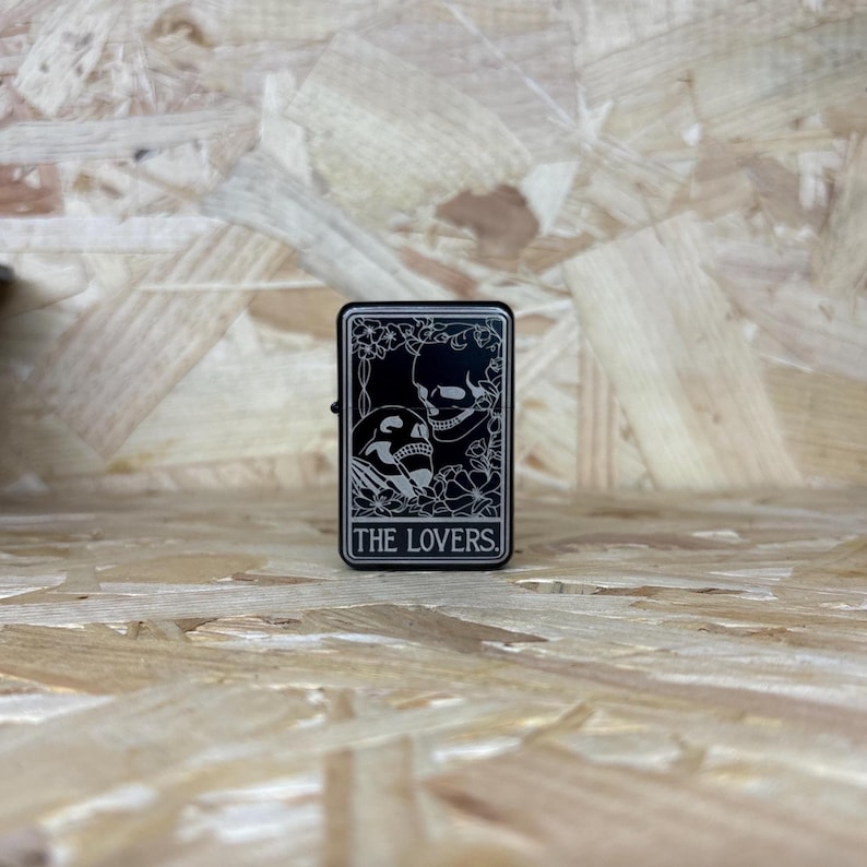 May include: Black metal lighter with a tarot card design. The card shows two skulls and floral details, with the text "THE LOVERS". The lighter is rectangular with a hinged lid.