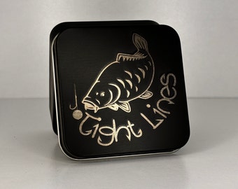May include: A black square tin with a gold carp illustration and the words "Tight Lines". The tin has a hinged lid and a silver rim. The design includes a fishing hook and a small round bait.