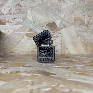 May include: Black metal lighter with a decorative design featuring skulls, flowers, and the text "THE LOVERS." The lighter is open, revealing the interior. The design is in a contrasting light color against the black background.