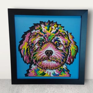 Könnte beinhalten: Gerahmtes Hundebild mit schwarzem Rahmen. Das Kunstwerk zeigt einen Hund mit einem lebendigen Fell aus Pink, Gelb, Grün und Blau vor einem hellblauen Hintergrund. Das quadratische gerahmte Kunstwerk wird auf einer hellgrauen Oberfläche präsentiert.