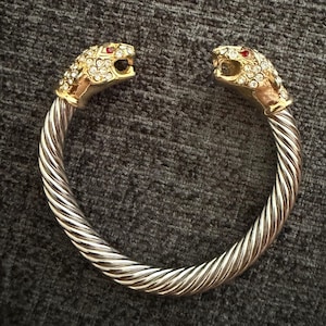 May include: A gold-tone cuff bracelet featuring two panther head terminals. Each head is encrusted with clear rhinestones and accented with a single red stone for the eye. The band consists of a twisted, cable-style metal design in a silver tone.