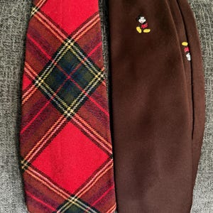 May include: Two neckties displayed side by side. The left tie features a red, green, and yellow plaid pattern. The right tie is solid brown and features a small, embroidered graphic of Mickey Mouse near the top center of the fabric.