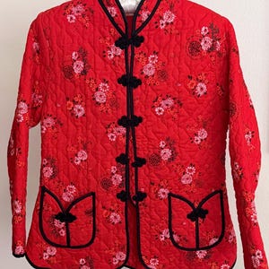 May include: A red quilted jacket featuring a floral pattern in shades of pink, white, and black. The jacket has a mandarin collar, long sleeves, and black piping trim. It closes with five black frog fasteners and includes two front patch pockets.