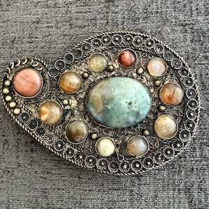 May include: An oval-shaped metal belt buckle with an intricate filigree border. The center features a large, smooth, oval-shaped green stone surrounded by smaller, round, polished stones in shades of peach, amber, and cream.