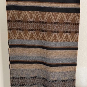 May include: A rectangular scarf featuring a woven pattern of horizontal stripes in black, gray, and brown. The design includes geometric diamond motifs and a fringed bottom edge. The word FENDI is embroidered in the lower right corner of the fabric.