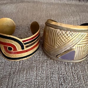 May include: Two wide cuff bracelets. The left cuff is gold-toned metal with black and red enamel swirl patterns. The right cuff is gold-toned metal featuring etched geometric lines, a braided border, and a single triangular purple inlay.