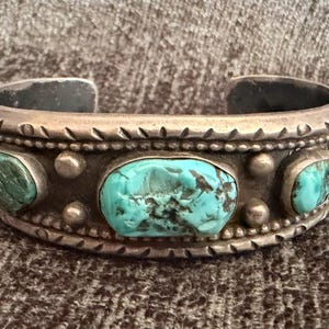 May include: A silver cuff bracelet featuring three turquoise stones set in a row. The metal band is decorated with a stamped pattern along the edges and small silver spheres placed between the stones. The center turquoise is the largest.