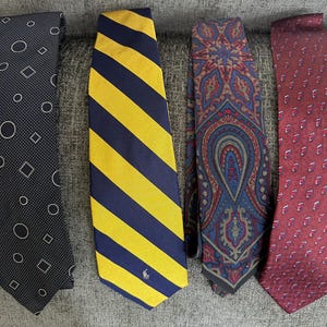 May include: Four neckties displayed side by side. The first is black with white geometric shapes. The second is yellow and navy blue striped with a small logo. The third features a multi-colored paisley pattern. The fourth is maroon with a small repeating pattern.
