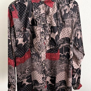 May include: A long-sleeved button-down shirt featuring a patchwork-style print in black, beige, and red. The design includes polka dots, marbled patterns, and abstract shapes. A vertical ruffled placket runs down the center front with small buttons.