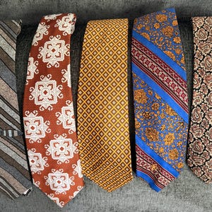 May include: Five neckties displayed side by side. The first features gray and brown diagonal stripes. The second is rust orange with white geometric patterns. The third is yellow with a small diamond grid. The fourth has blue, red, and floral motifs. The fifth is black with a tan paisley print.