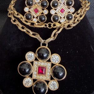 May include: A gold-tone necklace and matching clip-on earring set featuring a central square red stone surrounded by clear rhinestones, black cabochons, and a textured chain. The pendant and earrings share a symmetrical, multi-layered geometric design.