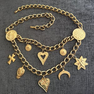 May include: A gold-tone chain belt featuring two circular medallions with embossed floral patterns and multiple dangling charms, including a cross, a heart, a star, a crescent moon, and two smaller circular discs with intricate detailing.