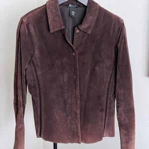 May include: A dark brown suede jacket featuring a classic collar, a front button closure, and long sleeves. The garment includes vertical decorative stitching details along the front panels and is lined with a dark fabric, hanging on a black hanger.