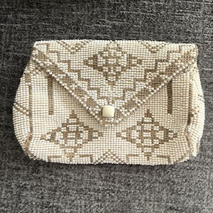 May include: A rectangular clutch bag covered in small white and metallic gold beads arranged in a geometric diamond pattern. The envelope-style flap features a single square white button closure at the center, securing the main compartment.